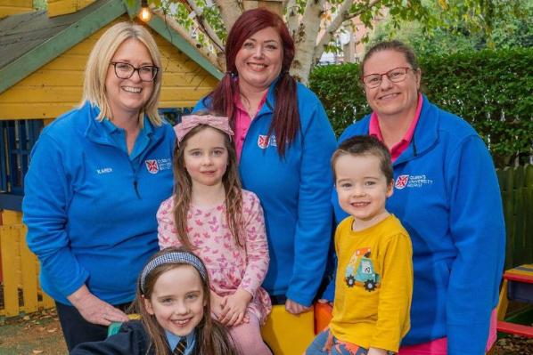Three QUB childcare staff in blue uniforms standing in front of three smiling children